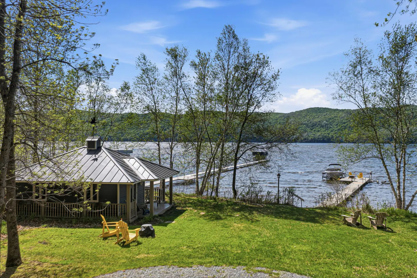 Lakefront cottage with a metal roof, a dock, and a boat on a sunny day. Two yellow chairs sit on the lawn.