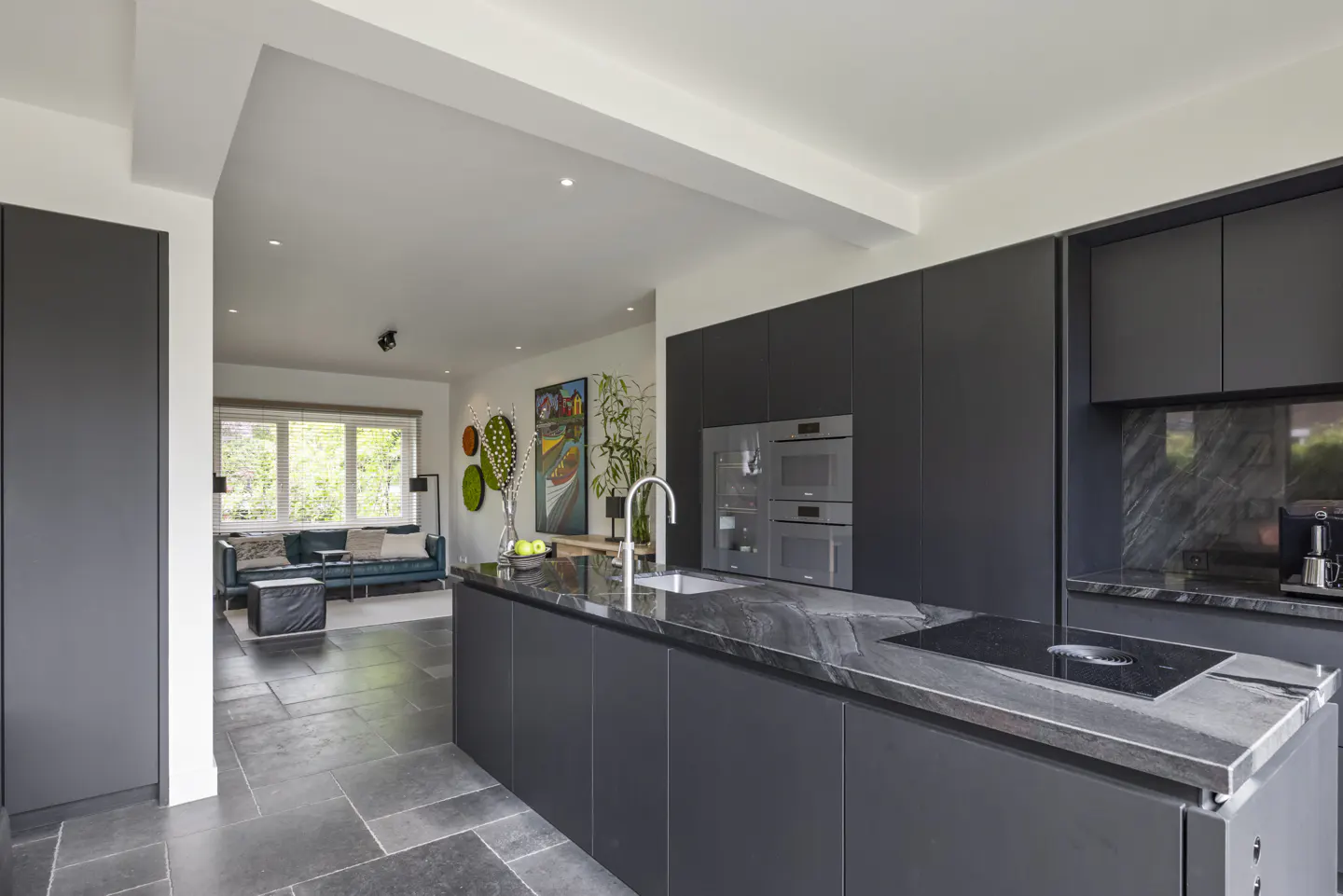 Open concept kitchen with dark cabinets, marble countertops, and stainless steel appliances. Living room with blue sofa in background.