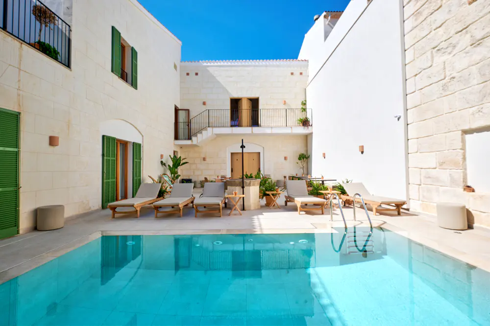 A turquoise pool is surrounded by lounge chairs in a courtyard with white stone buildings and green shutters.