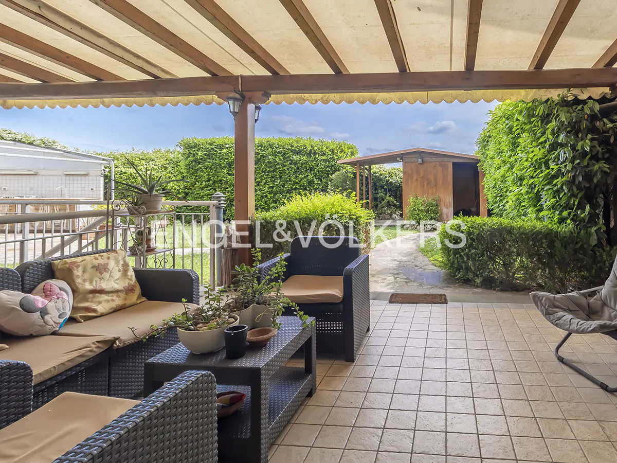 Covered patio with wicker furniture, beige cushions, and potted plants. A green hedge and a small shed are in the background.