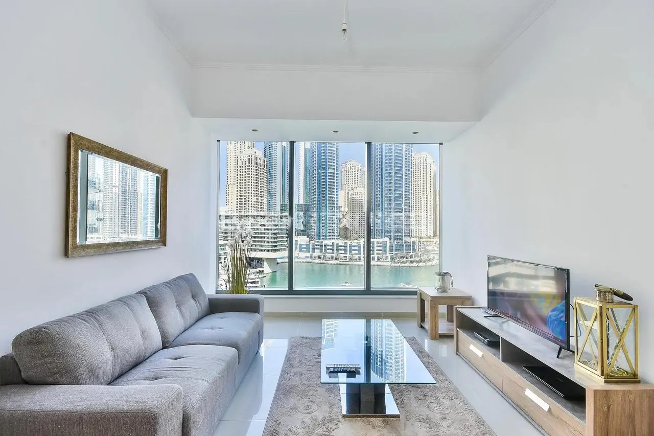 Bright living room with a gray sofa, glass table, and TV. Large window shows a city skyline with water views. Mirror on the wall.