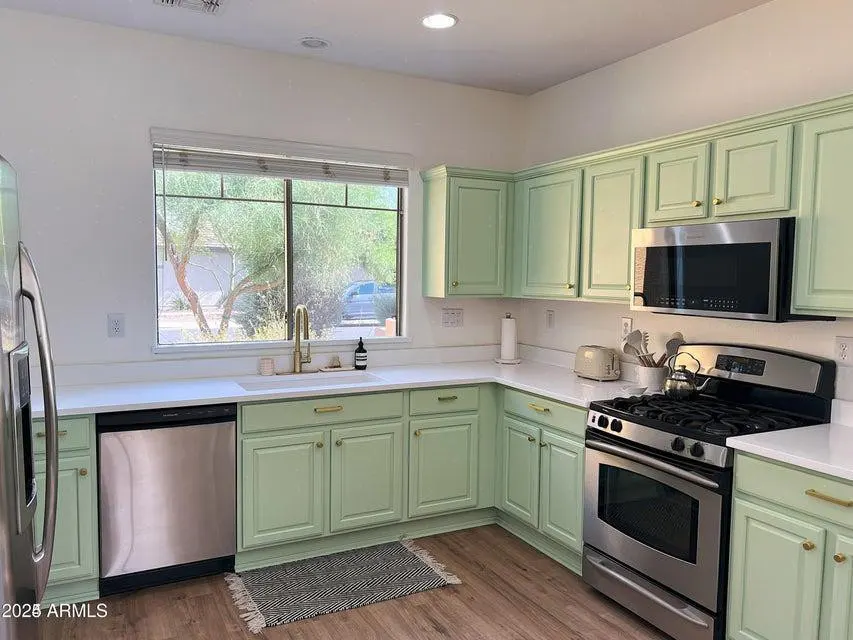 Bright kitchen with light green cabinets, stainless steel appliances, and white countertops. A window overlooks a tree.