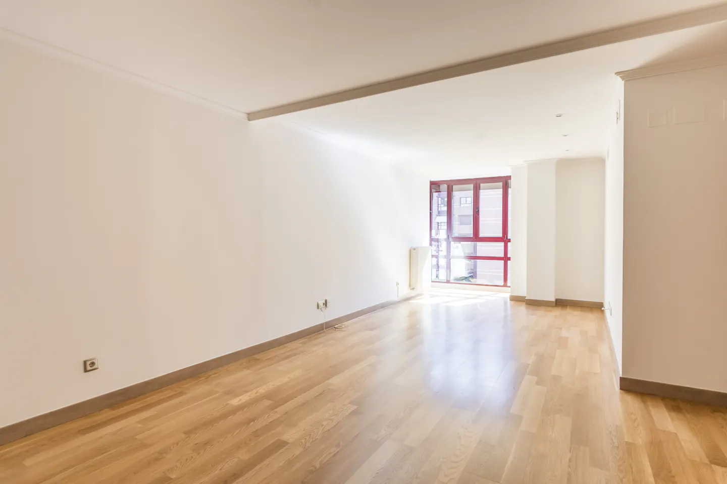 Bright, empty room with light wood floors and white walls. A red-framed window lets in sunlight.