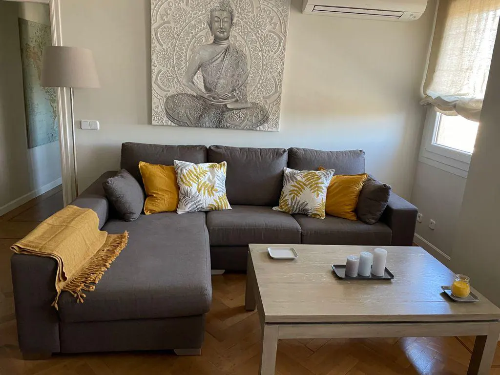 Living room with gray sectional sofa, yellow and leaf-patterned pillows, and a light wood coffee table with candles. A Buddha art piece hangs above the sofa.