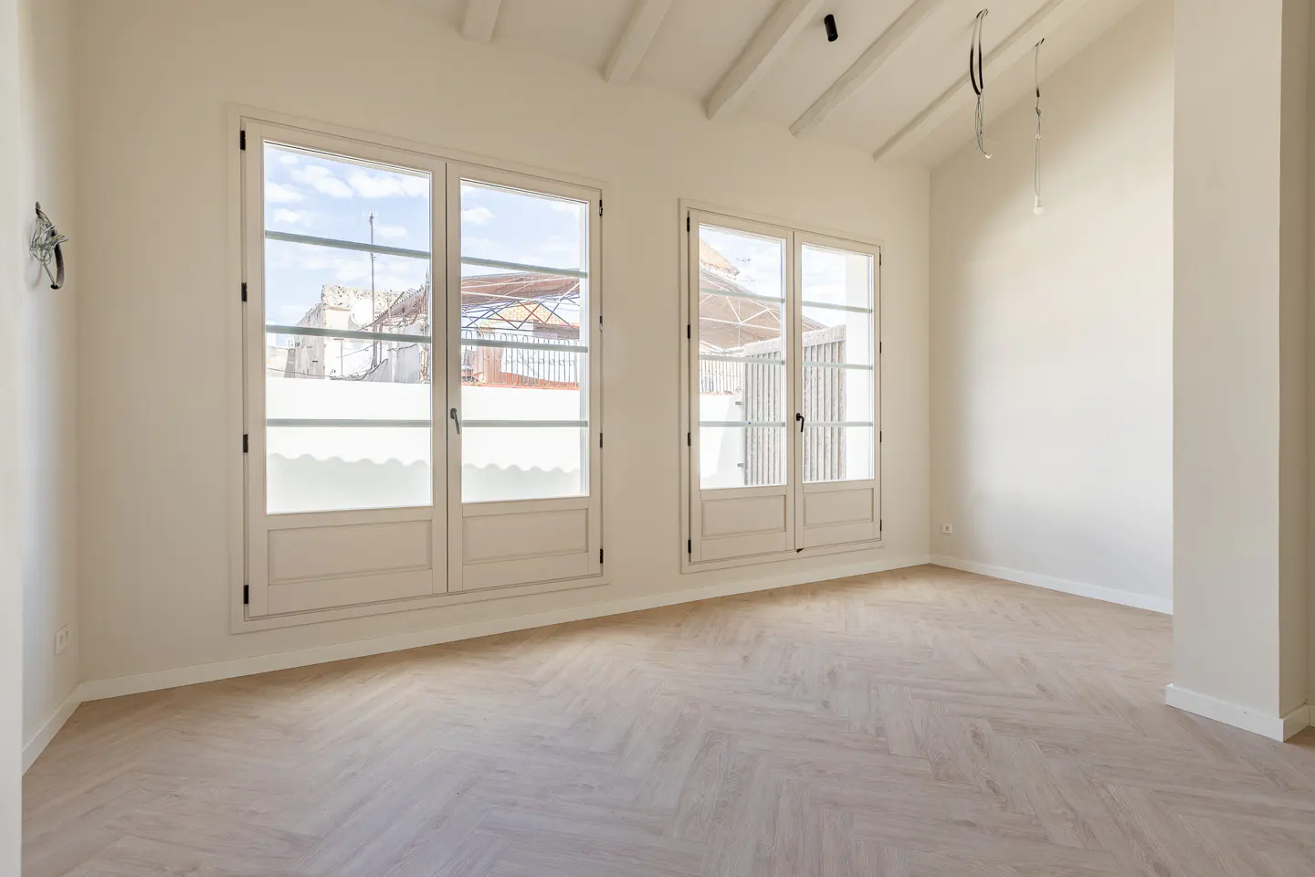 Bright, empty room with herringbone wood floors, white walls, and two large windows showing a building exterior.