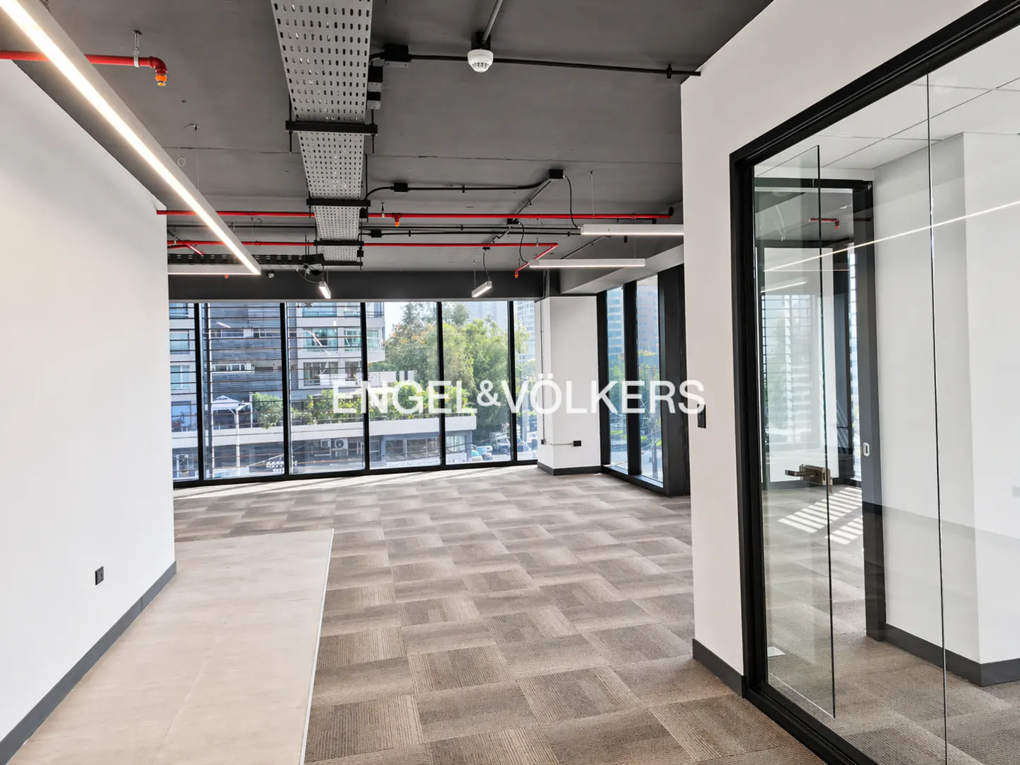An empty office space with large windows, gray carpet, and a glass-walled room. Red pipes and white cable trays are visible on the gray ceiling.