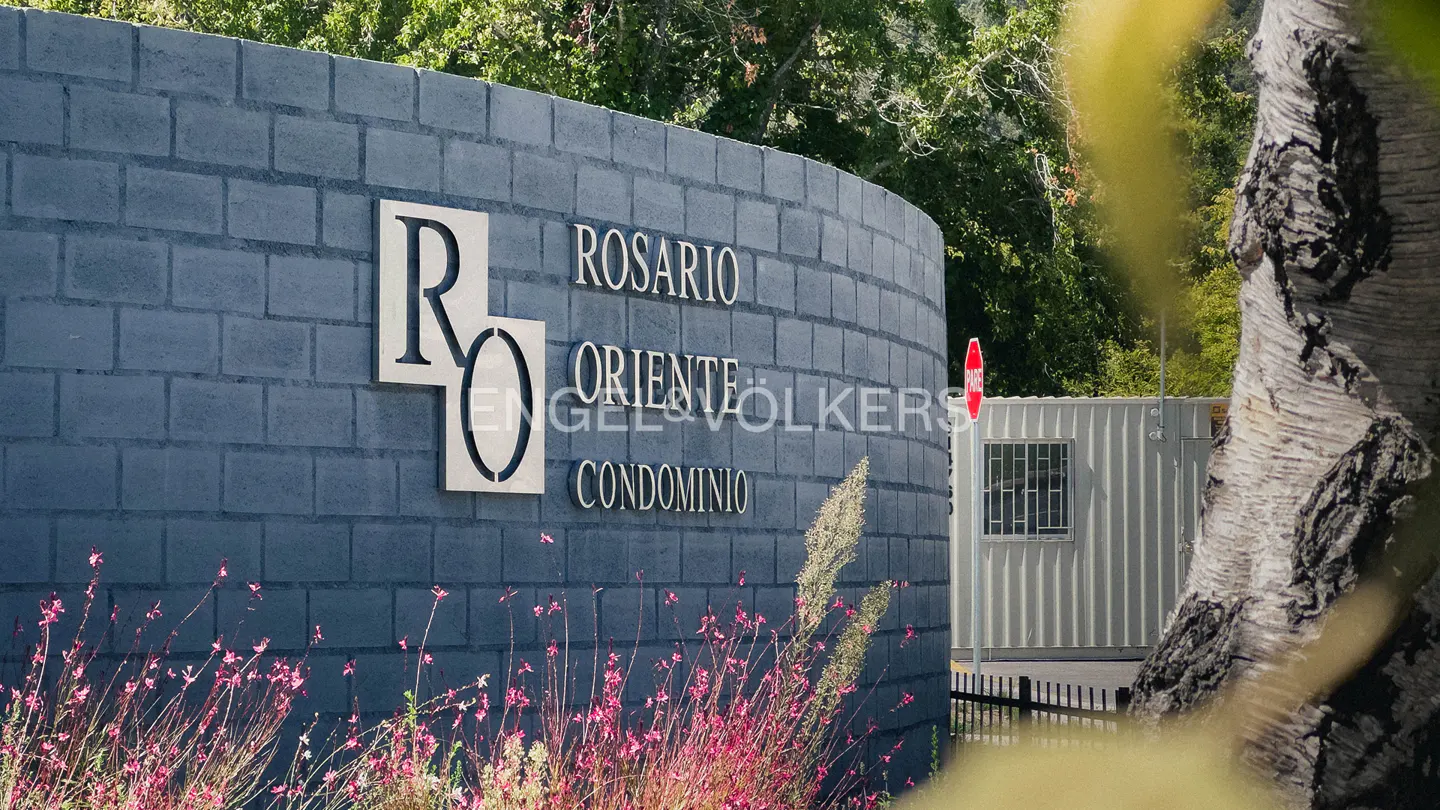 Entrance to Rosario Oriente Condominio with a gray brick wall and silver lettering. Pink flowers grow at the base. A white building is in the background.