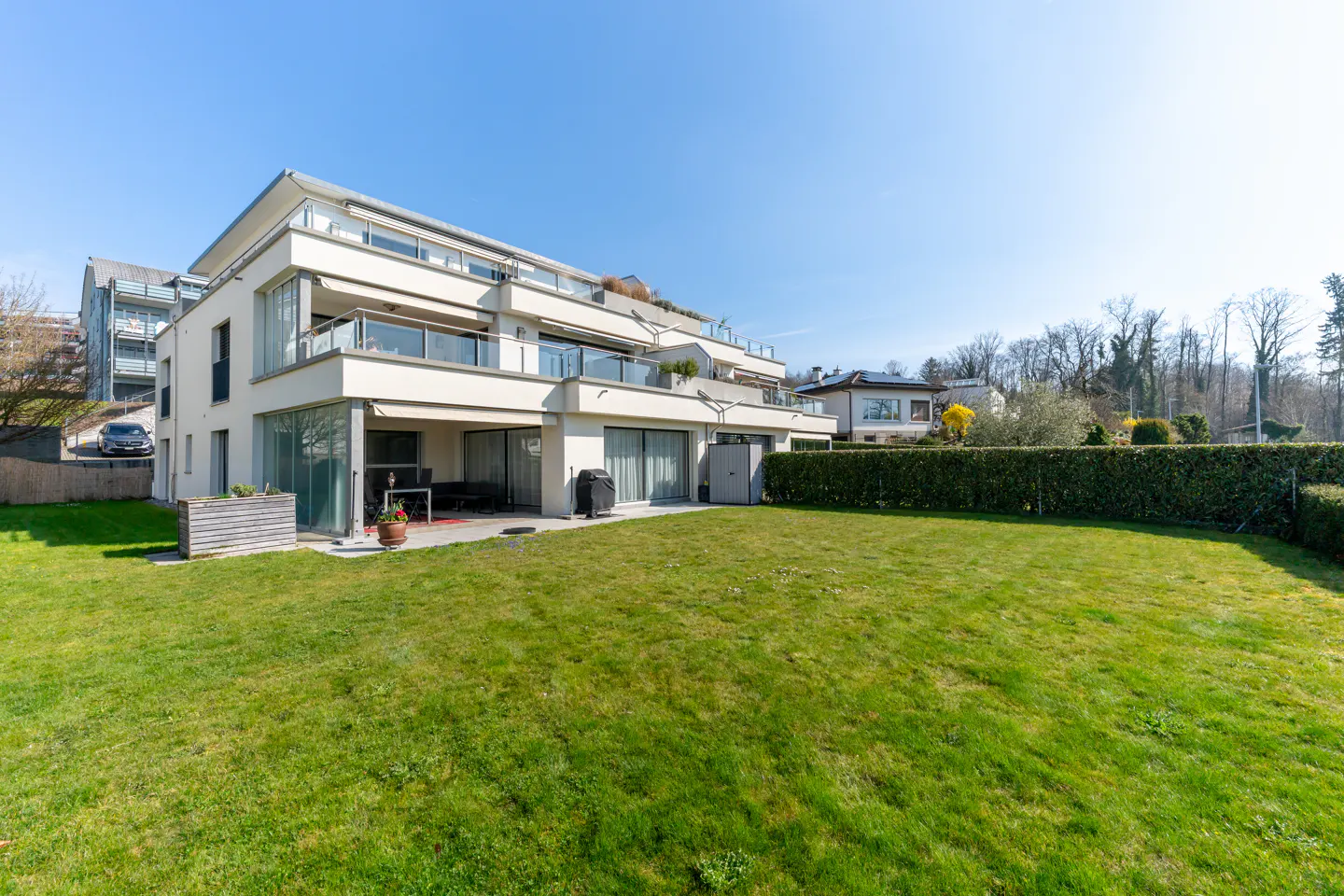 Modern white building with balconies and a large green lawn under a clear blue sky.