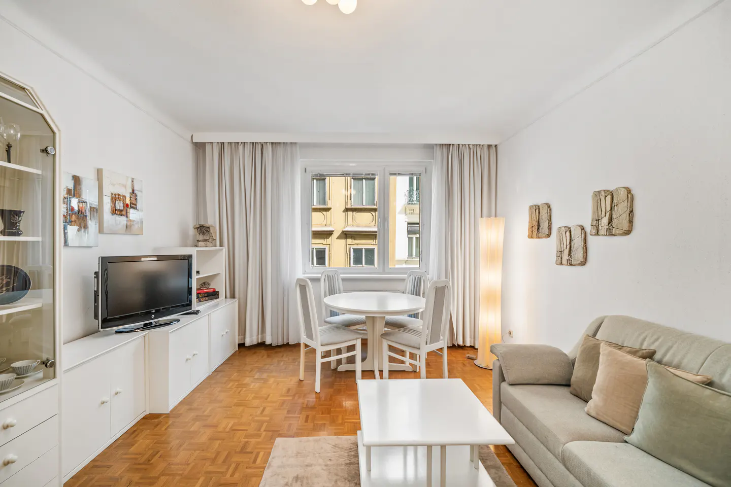 Bright living room with parquet floors, white walls, and a window view. A sofa, table, chairs, and built-in shelving unit furnish the space.