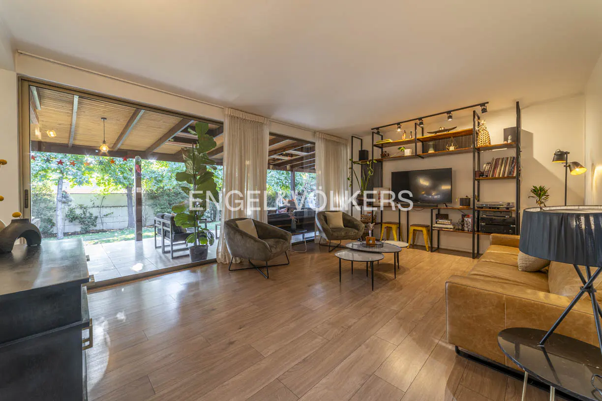 Living room with wood floors, tan sofa, two chairs, TV, and shelves. Large windows overlook a green backyard.