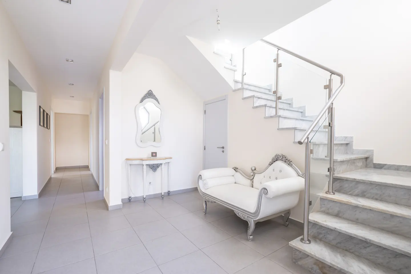 Bright foyer with marble stairs, glass railing, and white chaise lounge. A hallway leads to other rooms.