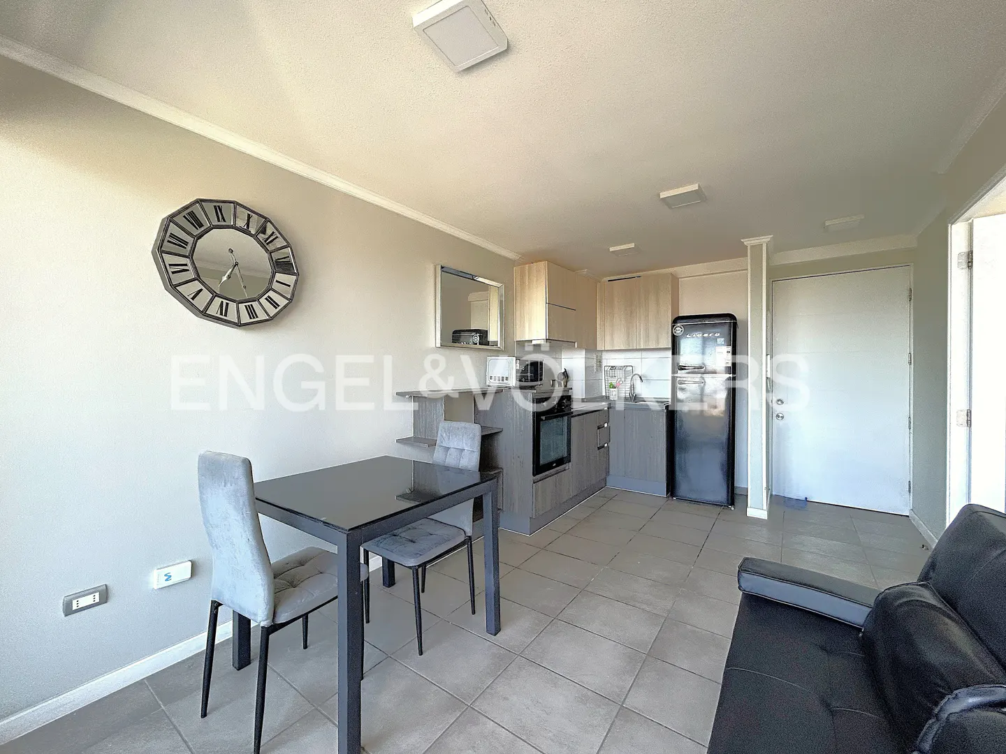A modern apartment interior with a black dining table, gray chairs, and a kitchen with a black refrigerator.