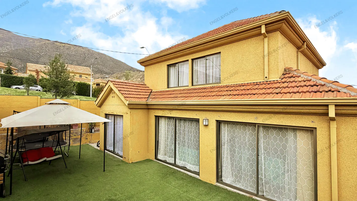 Backyard view of a two-story yellow house with a red tile roof and a mountain in the background. A white gazebo sits on the green lawn.