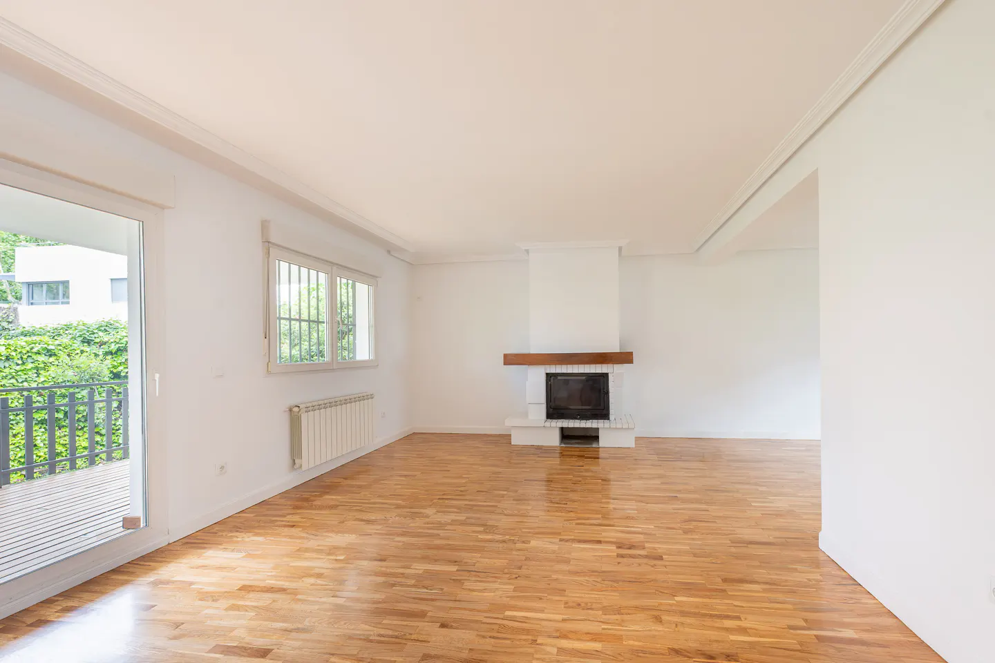 Bright, empty living room with hardwood floors, white walls, and a fireplace. A sliding glass door leads to a balcony with greenery.