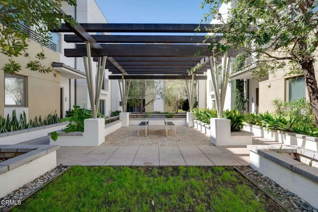 Courtyard view with a dark wood pergola over a brick patio with benches. Green grass and plants surround the patio. Buildings are in the background.