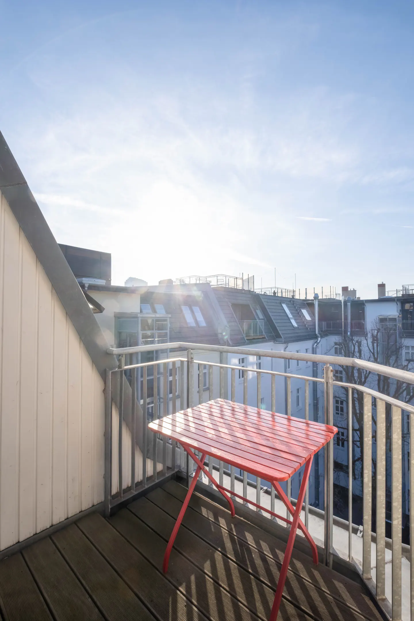 Balcony view with a red folding table on a wooden deck, metal railings, and city buildings under a bright blue sky.