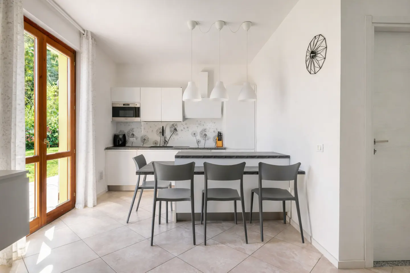 Bright, modern kitchen with white cabinets, gray chairs at a dark countertop island, and a wood-framed glass door to a green garden.