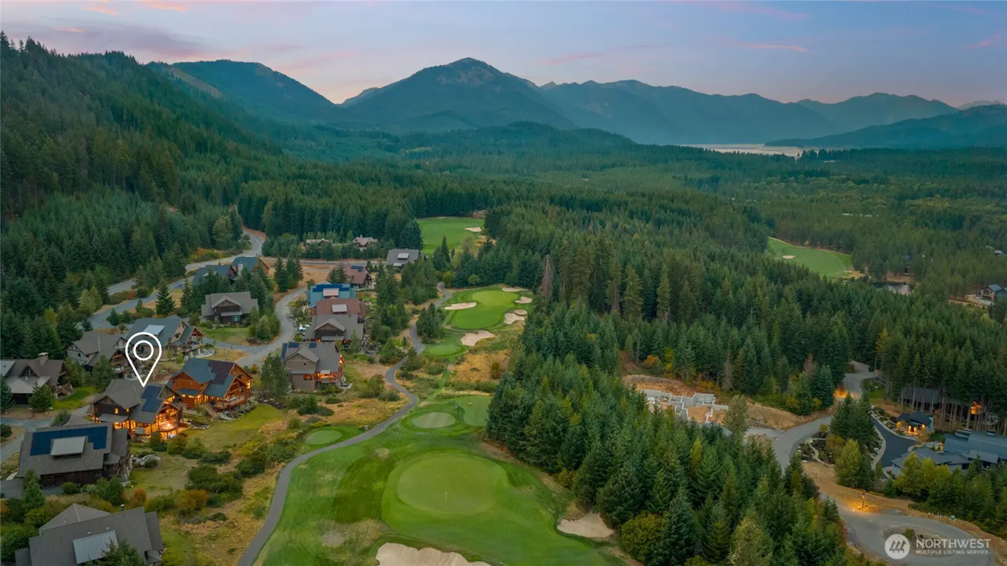 Aerial view of a mountain resort with a golf course, homes, and evergreen trees. Mountains are visible in the background.