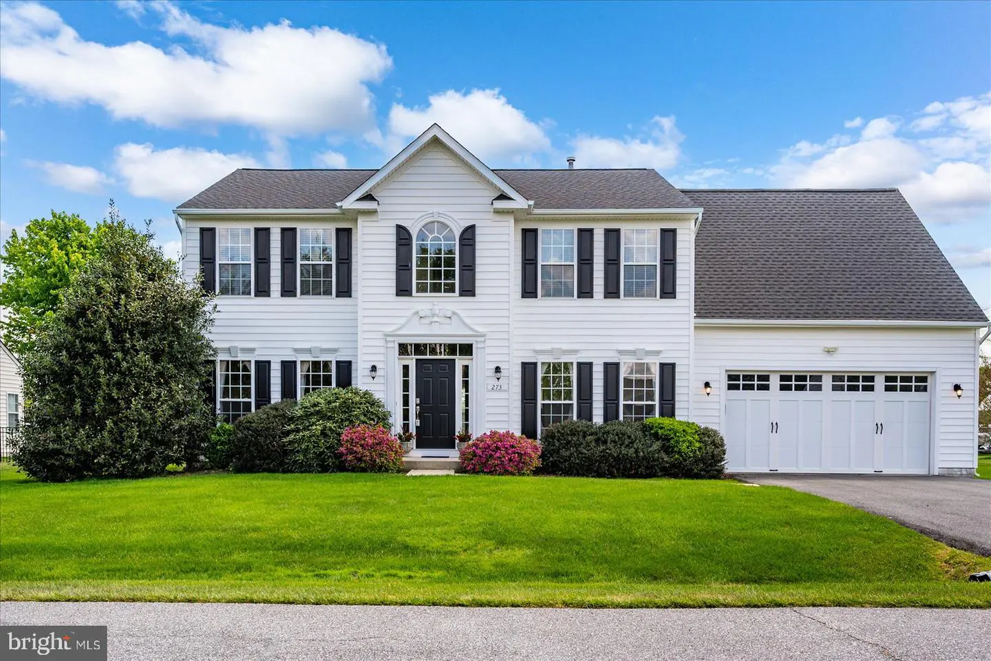 Two-story white house with black shutters, a black front door, and a two-car garage on a green lawn under a blue sky.