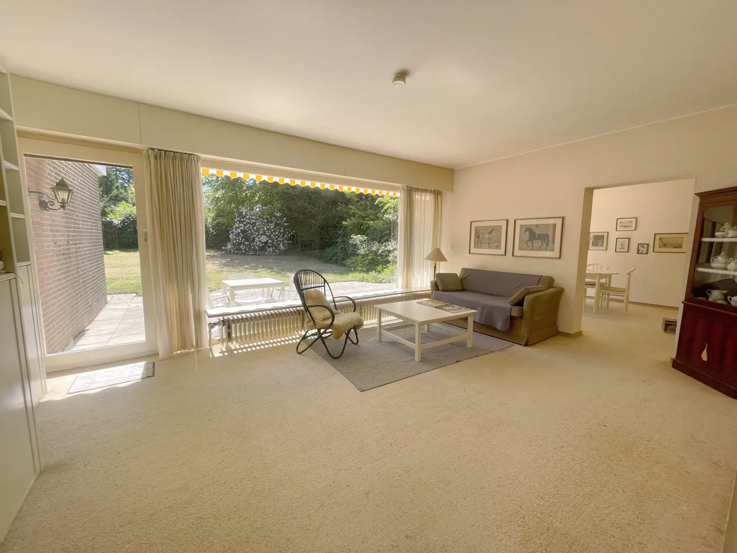 Living room with beige carpet, sofa, chair, and white coffee table. Large window overlooks a green garden with a white table.