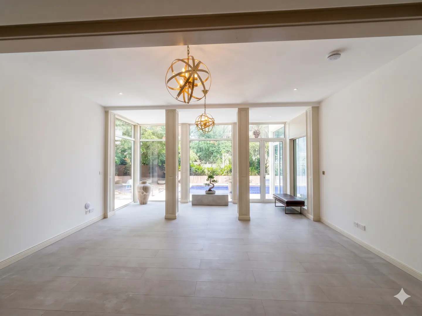 Bright, minimalist room with light tile floors, two gold orb chandeliers, and floor-to-ceiling windows overlooking a pool.
