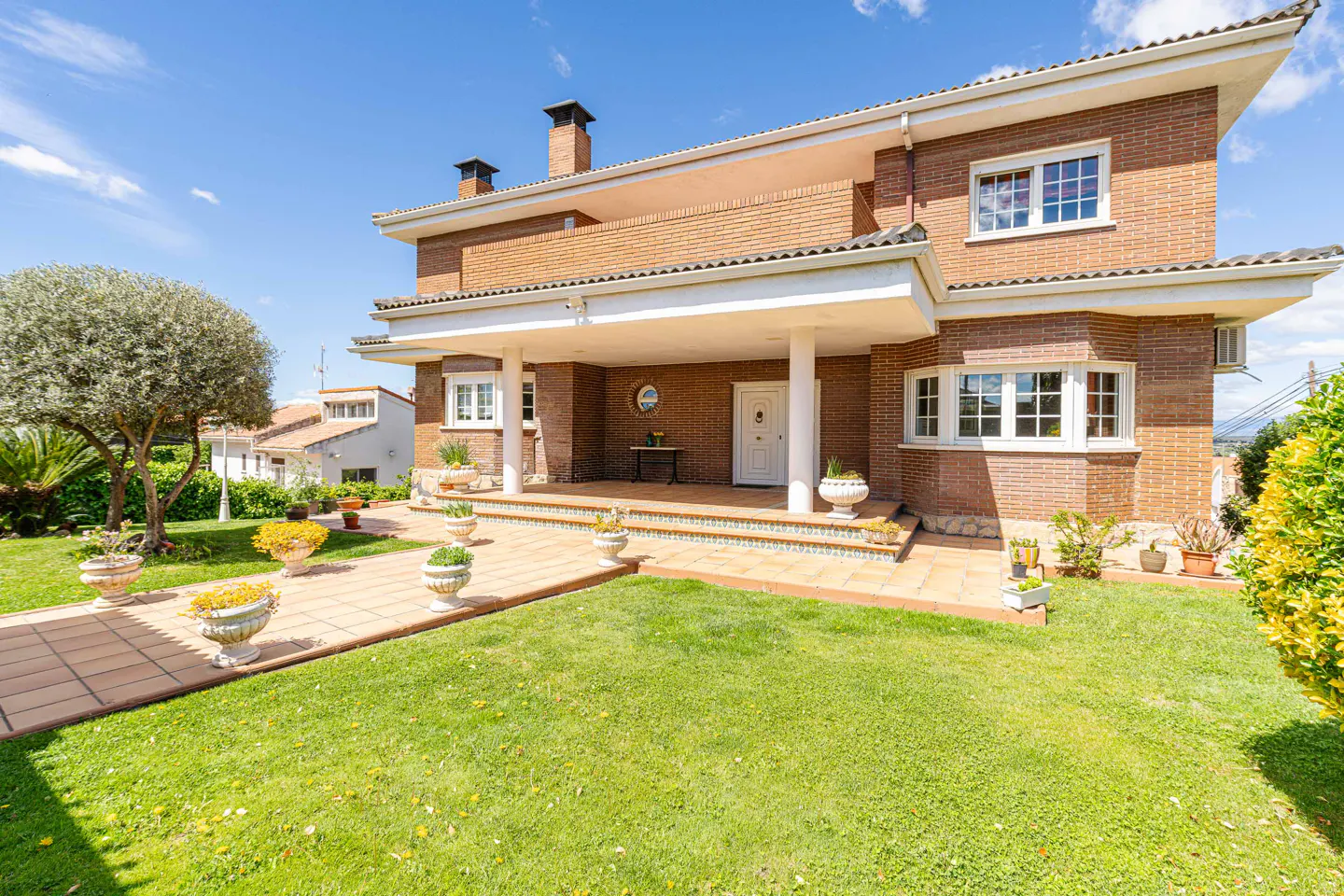 Two-story brick house with a white porch, green lawn, and tiled walkway under a blue sky.