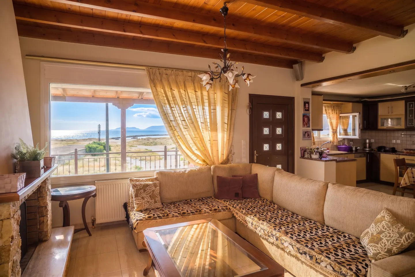 Living room with a large window view of the ocean. A tan sectional sofa with patterned cushions faces a glass coffee table. A kitchen is visible in the background.