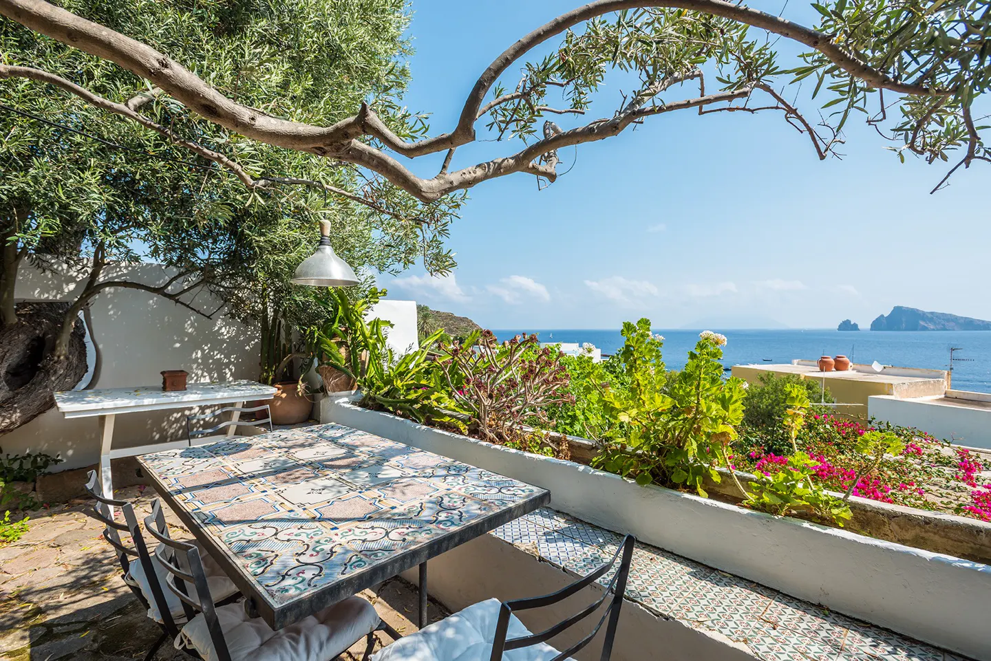 Outdoor patio with a patterned table, chairs, and a view of the ocean and island under a tree.