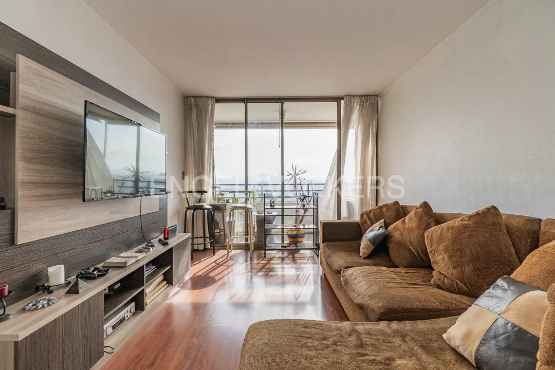 Living room with brown sectional sofa, TV, and balcony with city view. Hardwood floors and neutral walls.
