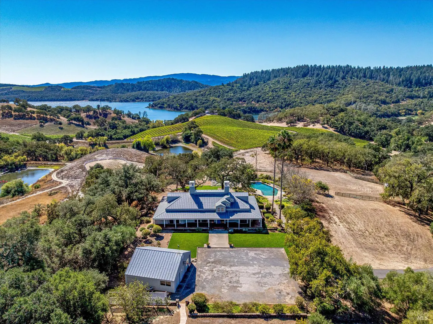 Aerial view of a large, light-colored house with a pool, vineyard, and lake in the background on a sunny day.