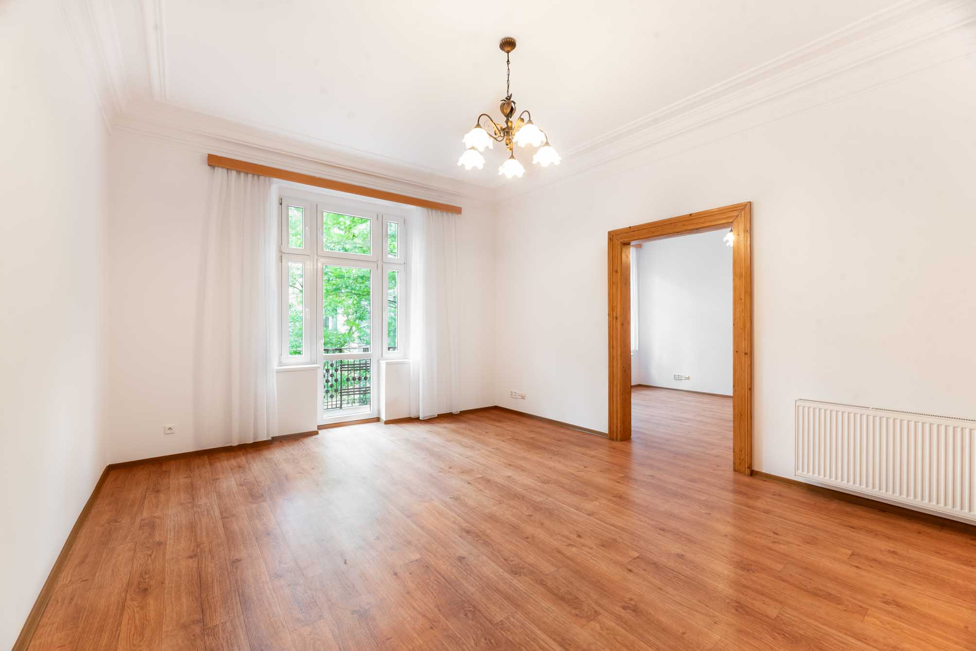 Bright, empty room with wood floors, white walls, a classic chandelier, and a balcony door overlooking green trees.