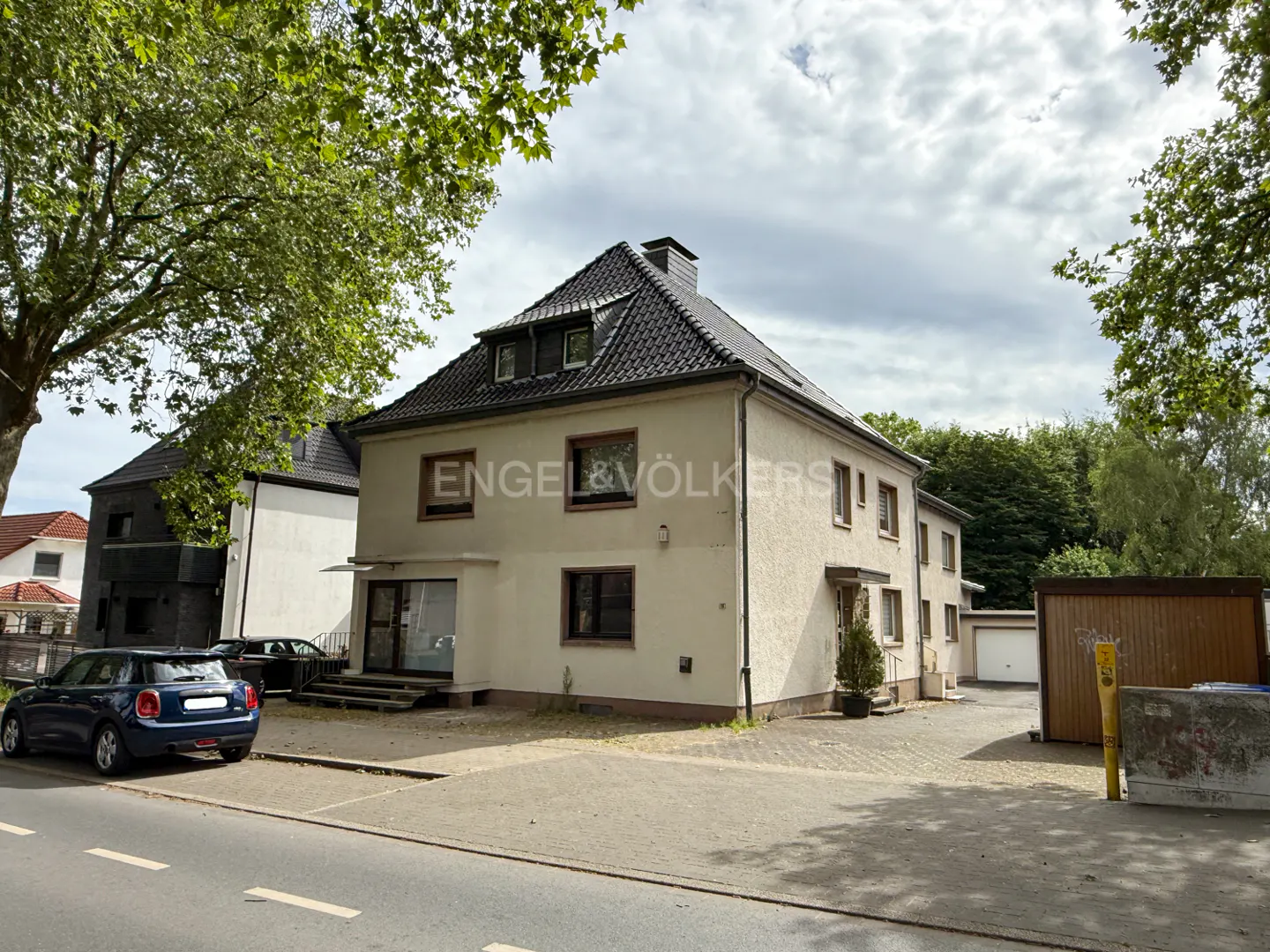 A two-story beige house with a dark tiled roof, a blue car parked in front, and a wooden garage on a sunny day.
