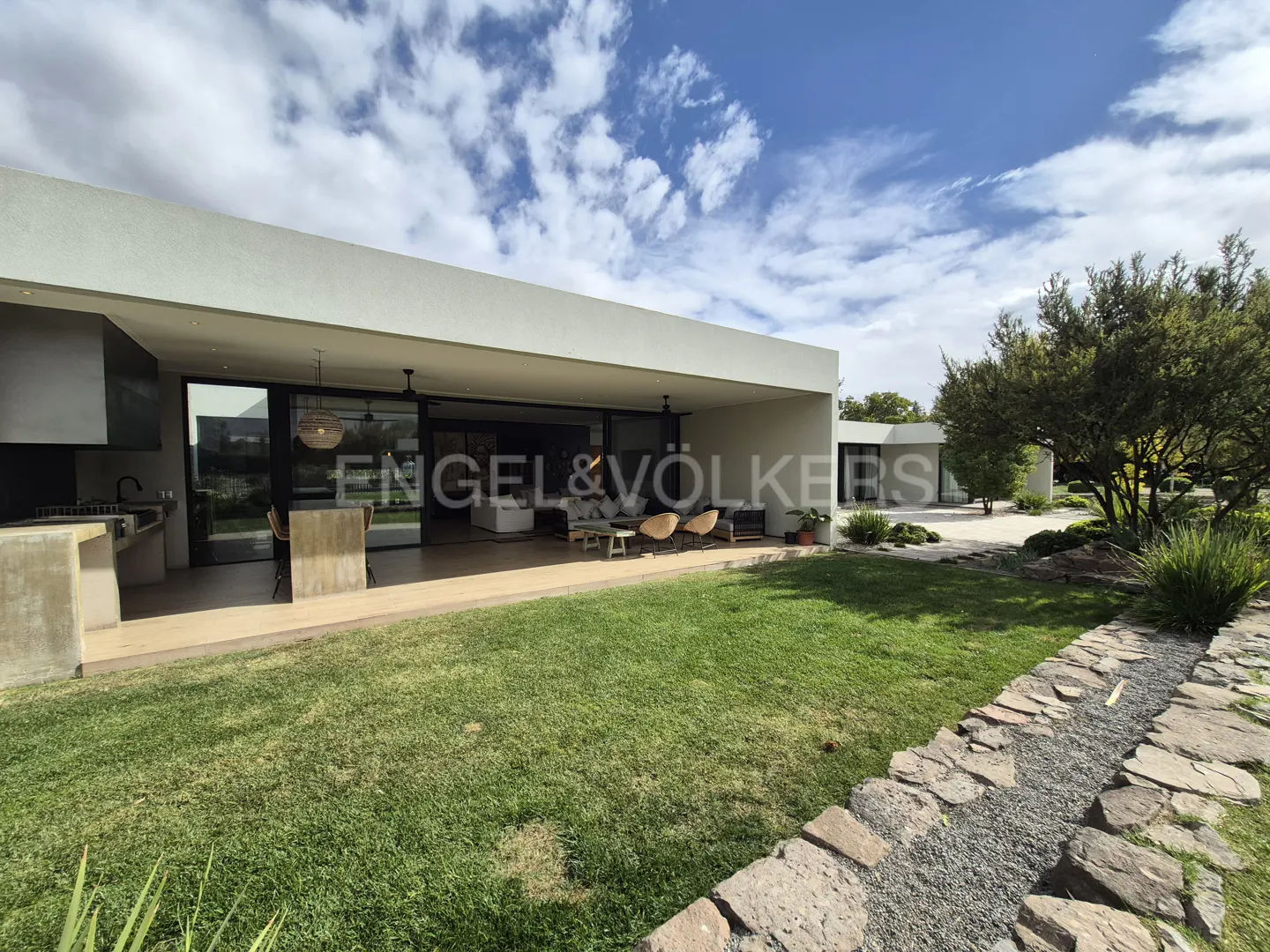 Modern white house with a covered patio, outdoor kitchen, and green lawn under a blue sky with clouds.