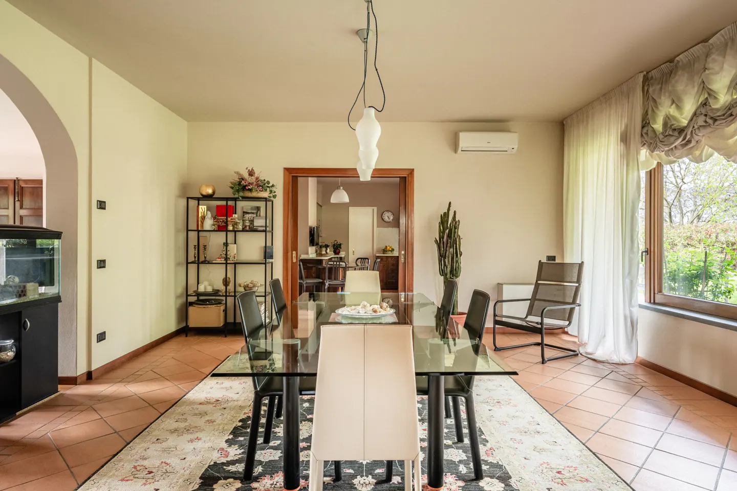 Dining room with glass table, black chairs, and beige walls. A black shelf and aquarium are on the left, and a window with curtains is on the right.