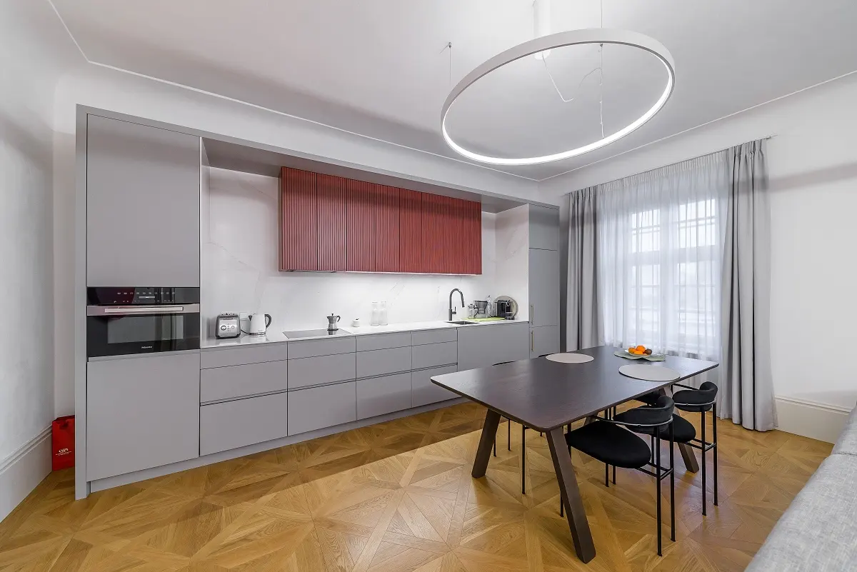 A modern kitchen with gray cabinets, a red accent cabinet, and a dark wood table with black chairs. A circular light fixture hangs above.