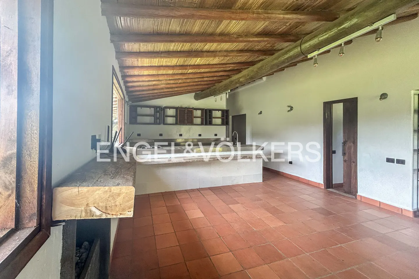 A spacious room with terracotta tile flooring, a wooden ceiling, and a kitchen area with white countertops.