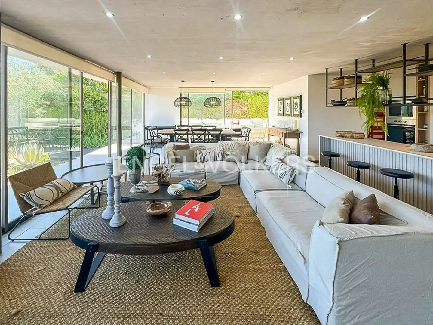 Open-concept living space with a white sectional sofa, round coffee tables, and floor-to-ceiling windows overlooking a lush green garden.