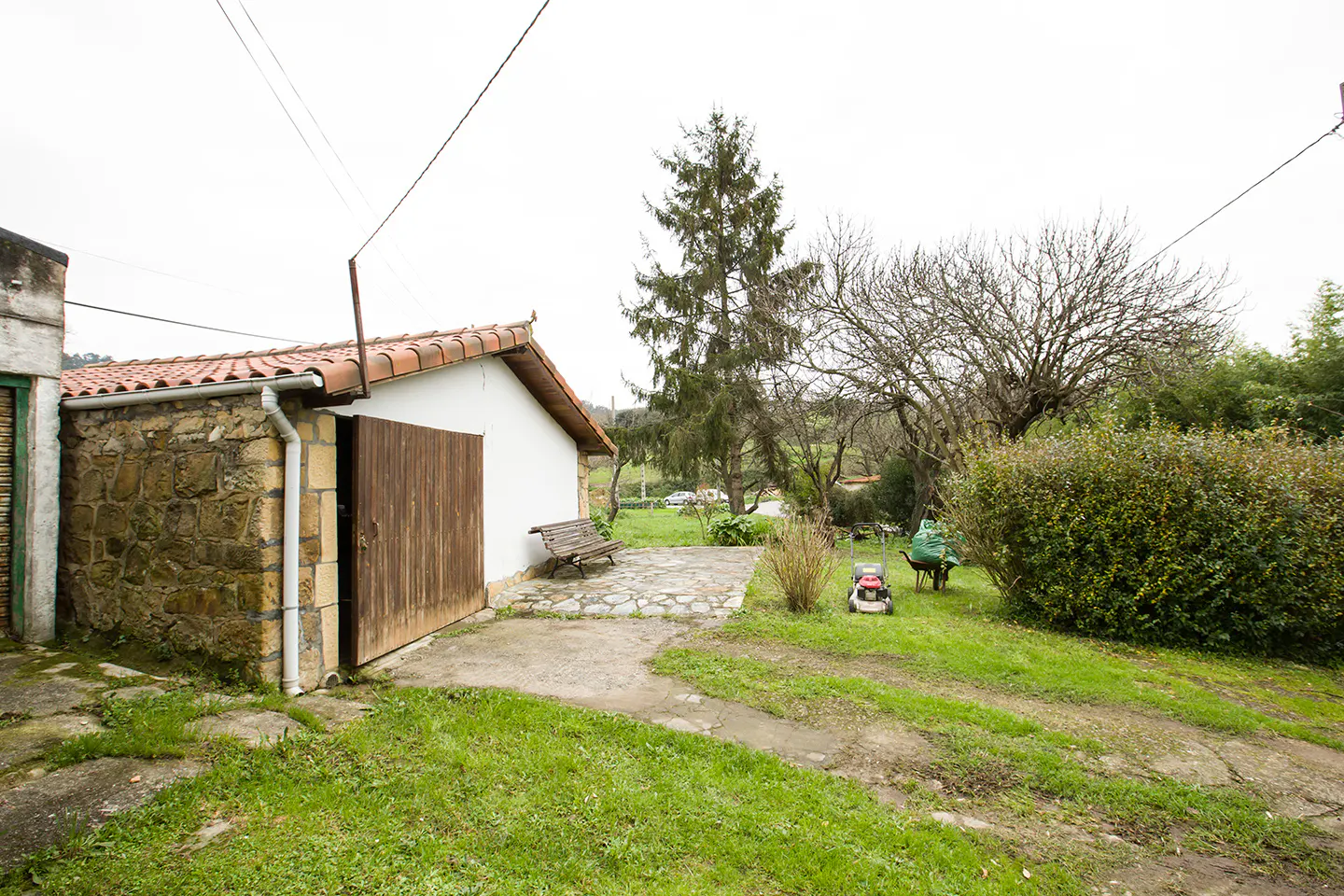 Exterior view of a stone and white building with a red tile roof, a wooden gate, and a green lawn. A bench sits nearby.