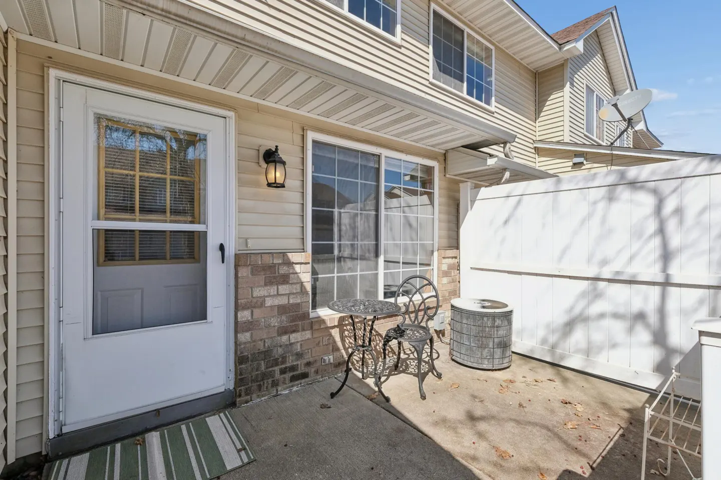 Exterior view of a tan two-story townhouse with a white door, sliding glass door, and patio furniture.