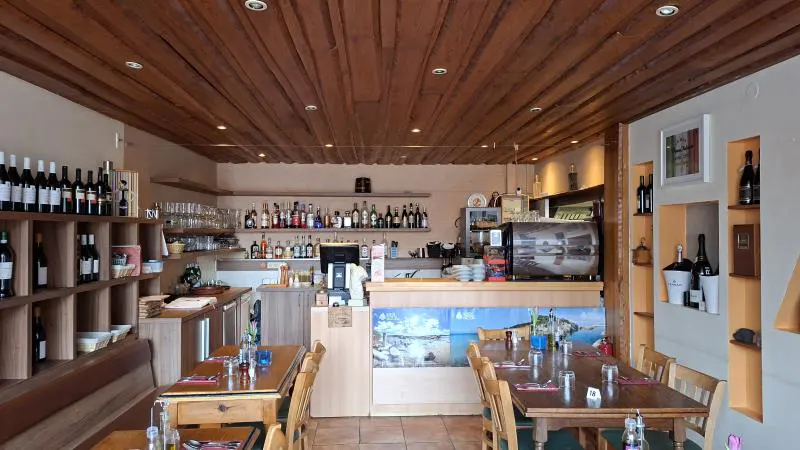 Restaurant interior with wooden ceiling, tables set with glasses and cutlery, and shelves stocked with wine bottles.