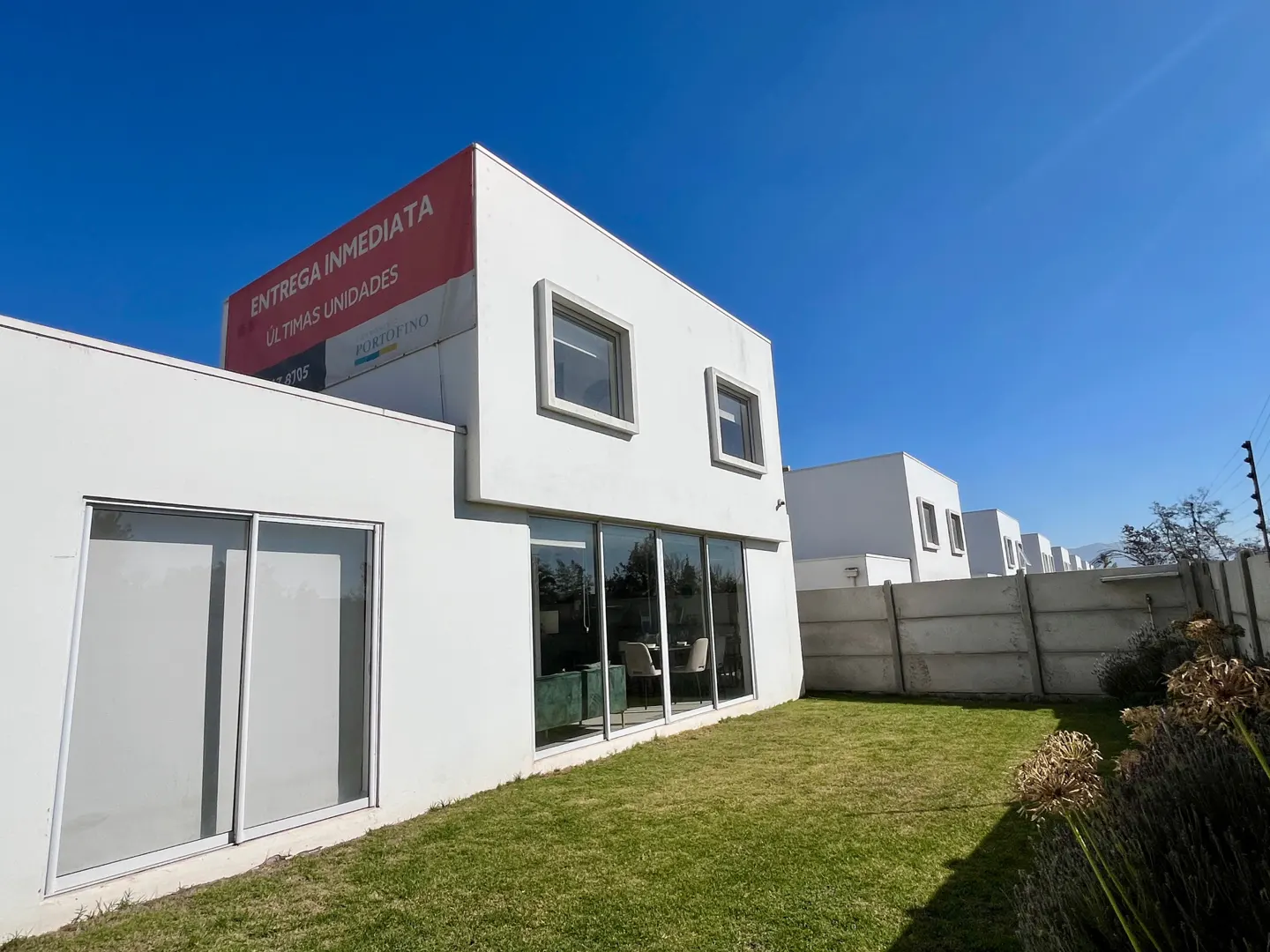 Modern white house with a green lawn under a blue sky. A red sign reads "Entrega Inmediata". Interior furniture is visible through large windows.
