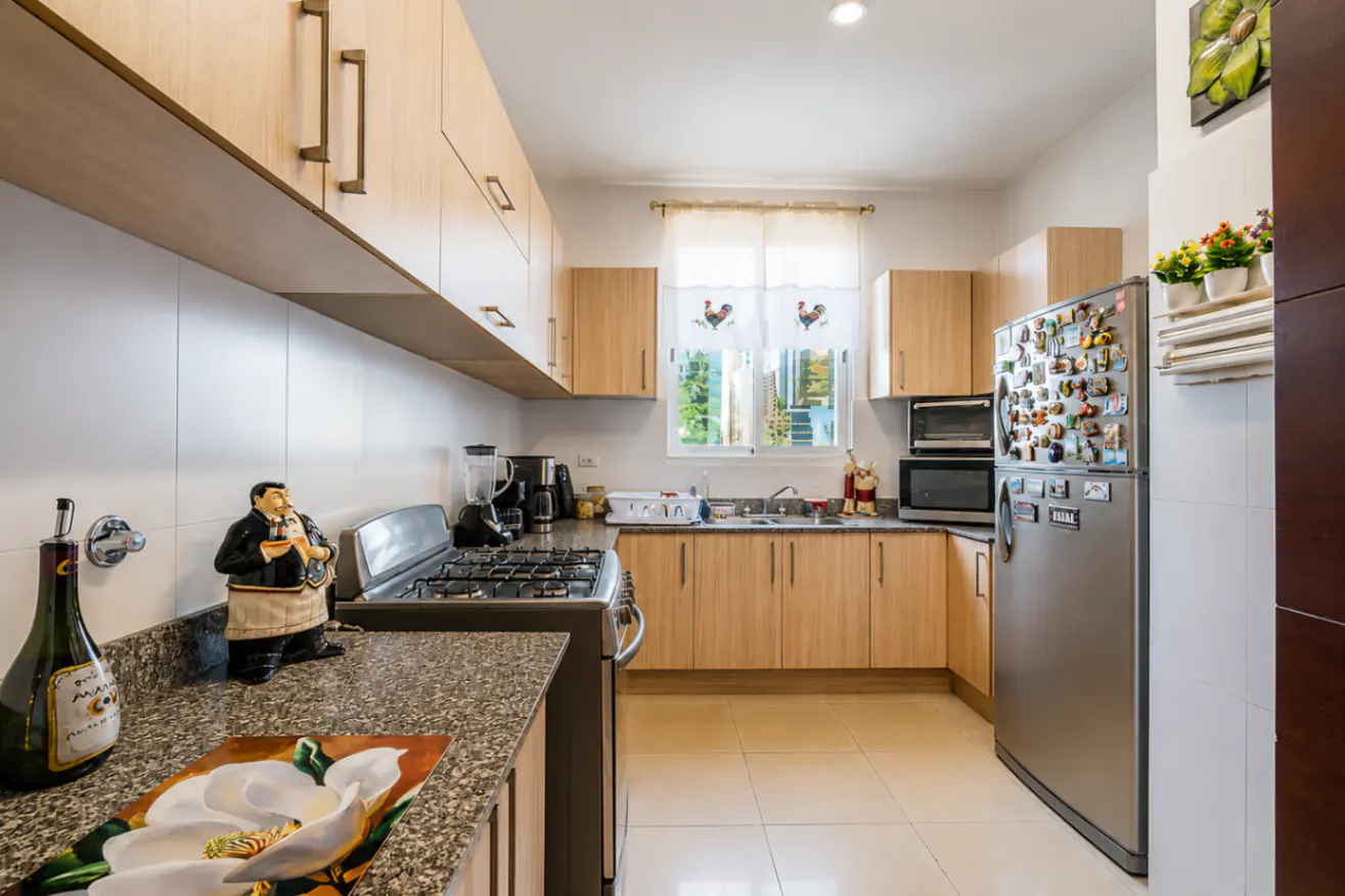Bright kitchen with light wood cabinets, stainless steel appliances, and granite countertops. A window with rooster curtains lets in natural light.