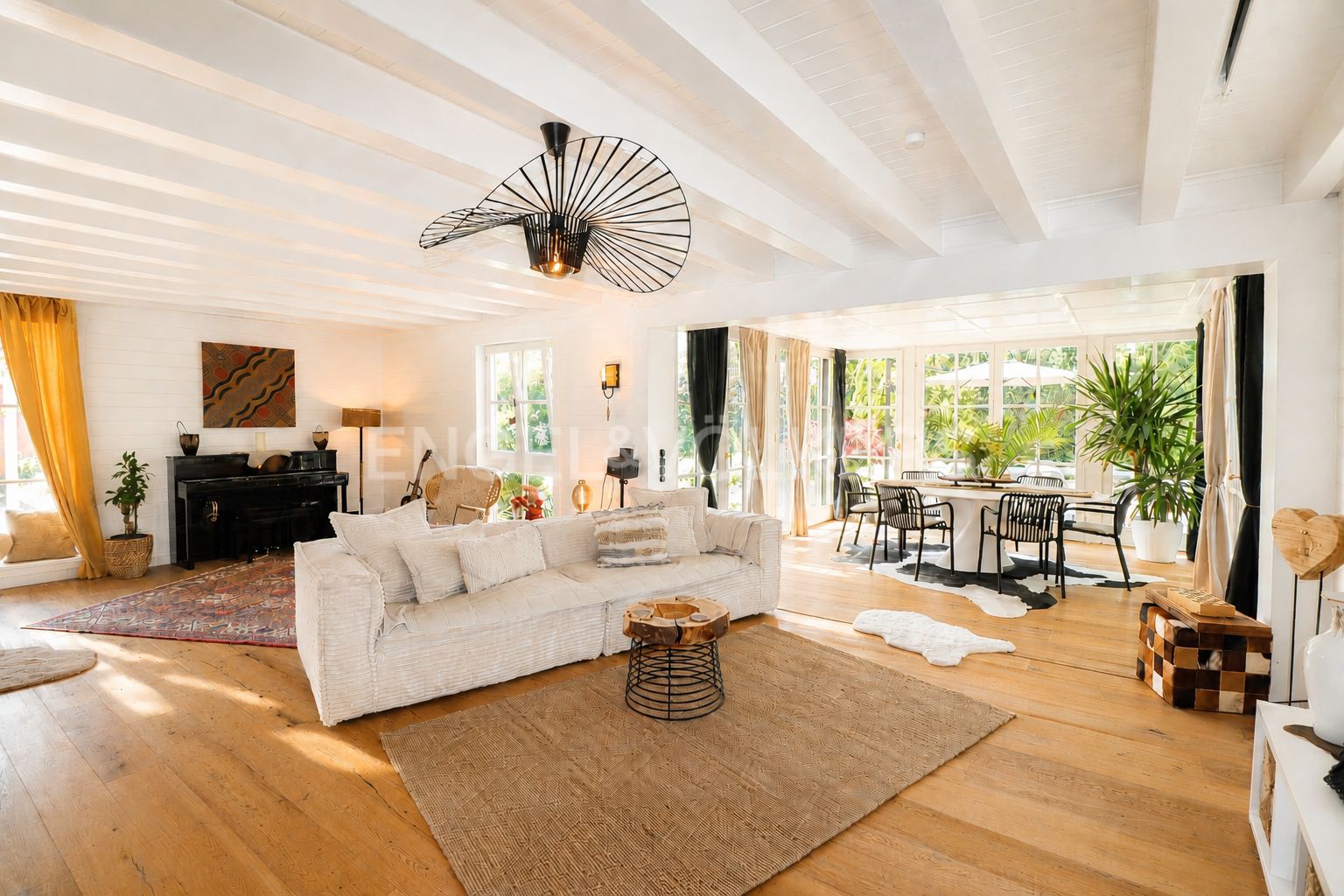 Bright living room with white sofa, wooden floors, exposed beam ceiling, modern fan, and dining area with large windows.