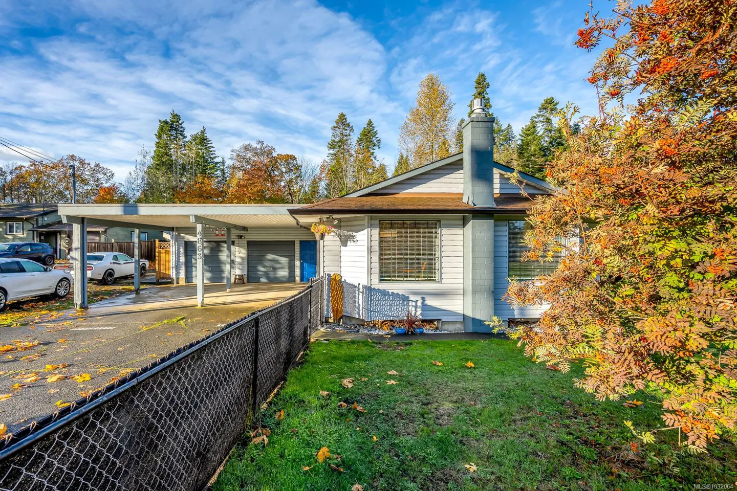 A single-story light blue house with a carport, a black chain link fence, and fall foliage.