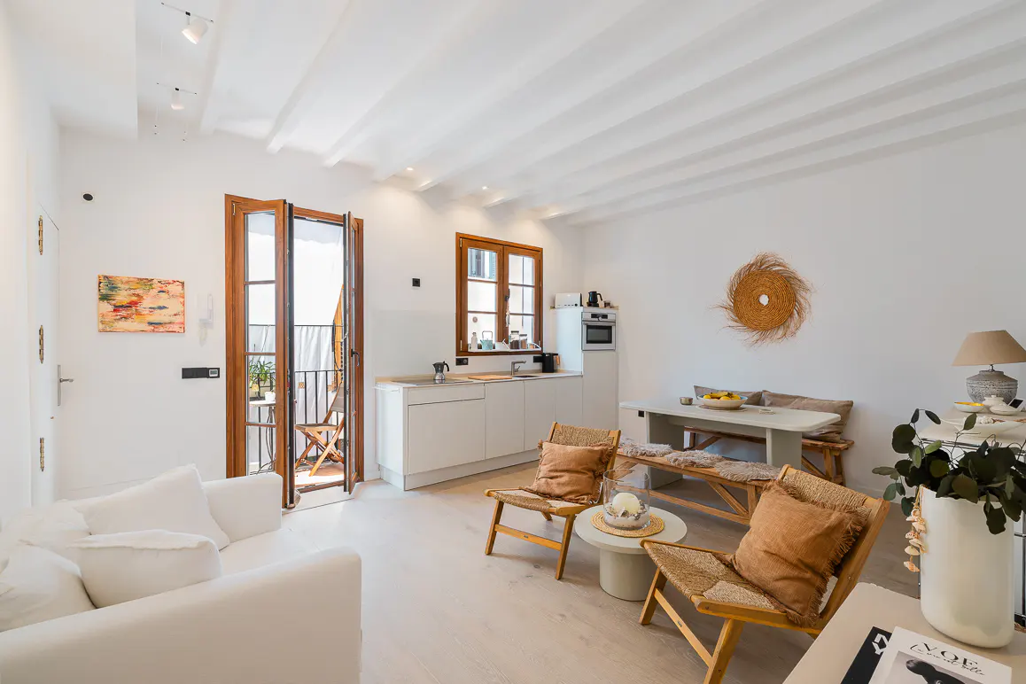 Bright, airy living space with white walls, wood accents, and a balcony. A white sofa and woven chairs create a cozy atmosphere.