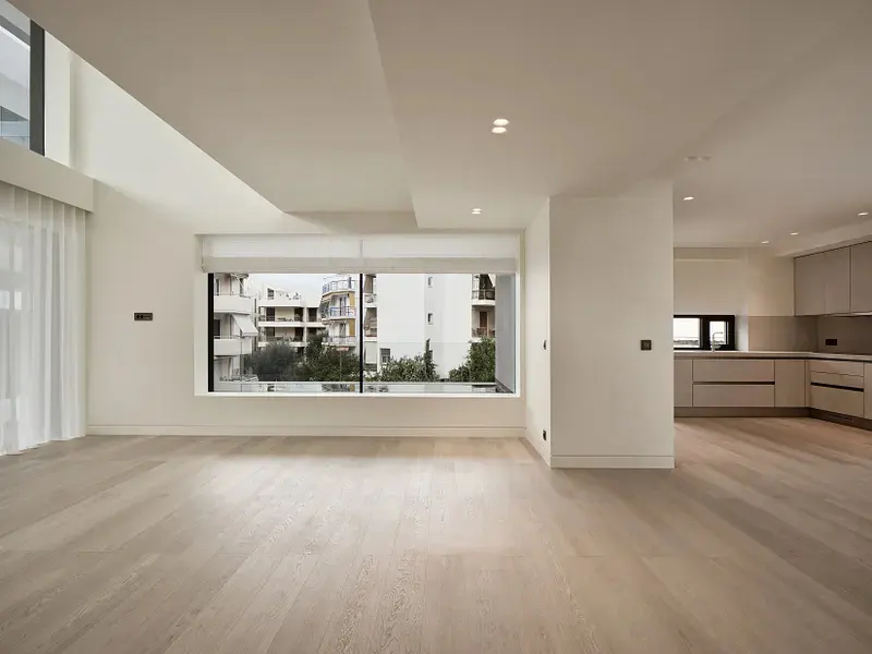 Bright, empty living room with light wood floors and white walls. Large window overlooks city buildings. Kitchen visible in the background.