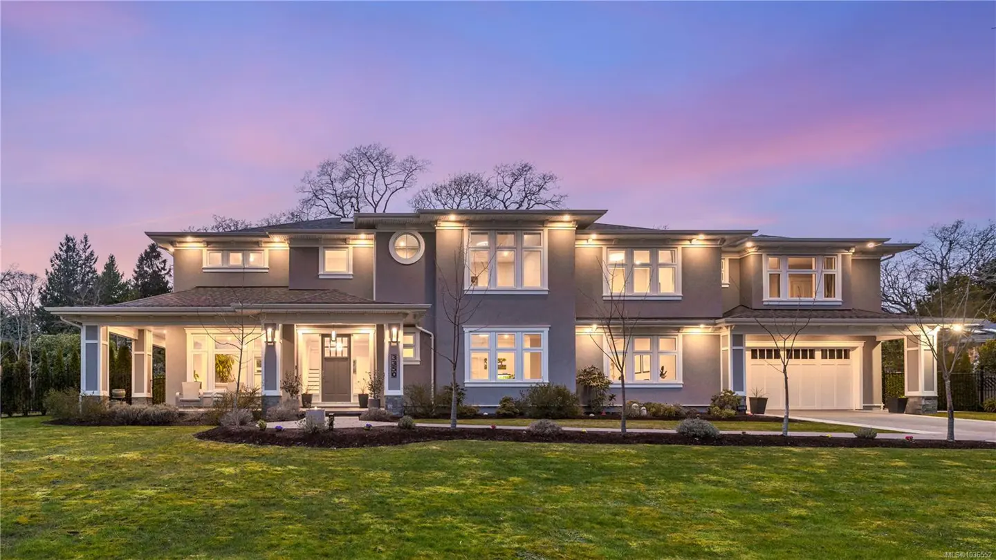 Two-story gray house with white trim, illuminated by outdoor lights, sits on a green lawn under a pink and blue twilight sky.