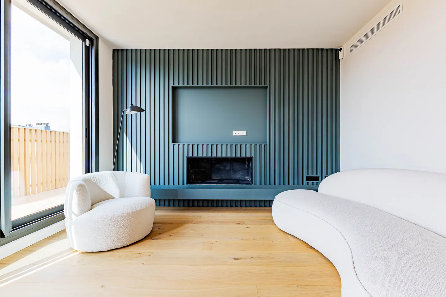Bright living room with wood floors, a blue accent wall with a fireplace, and white curved furniture. A large window lets in natural light.