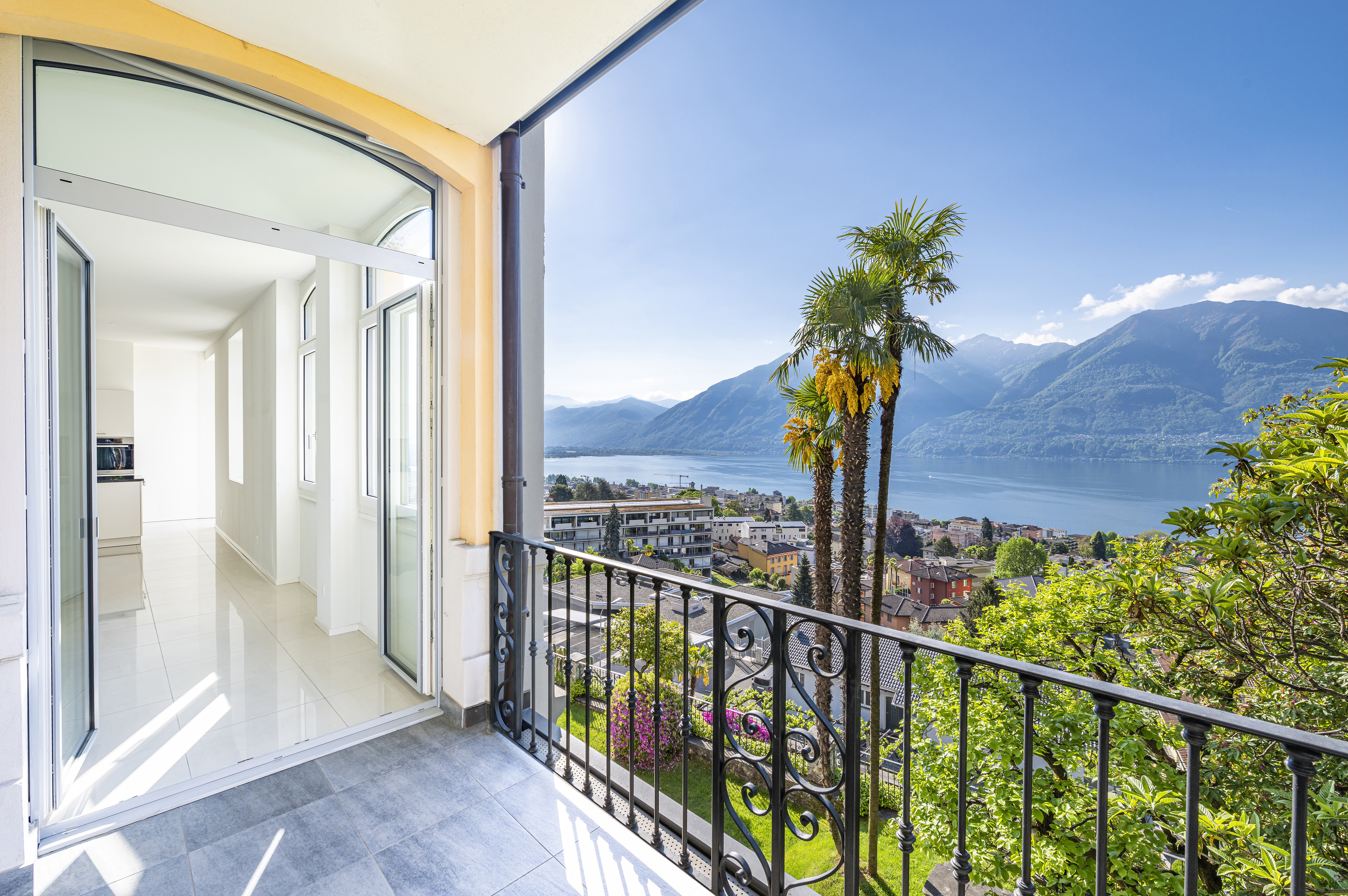 Balcony view of a lake and mountains. Black iron railing, palm trees, and a glimpse of a white interior with tile floors.