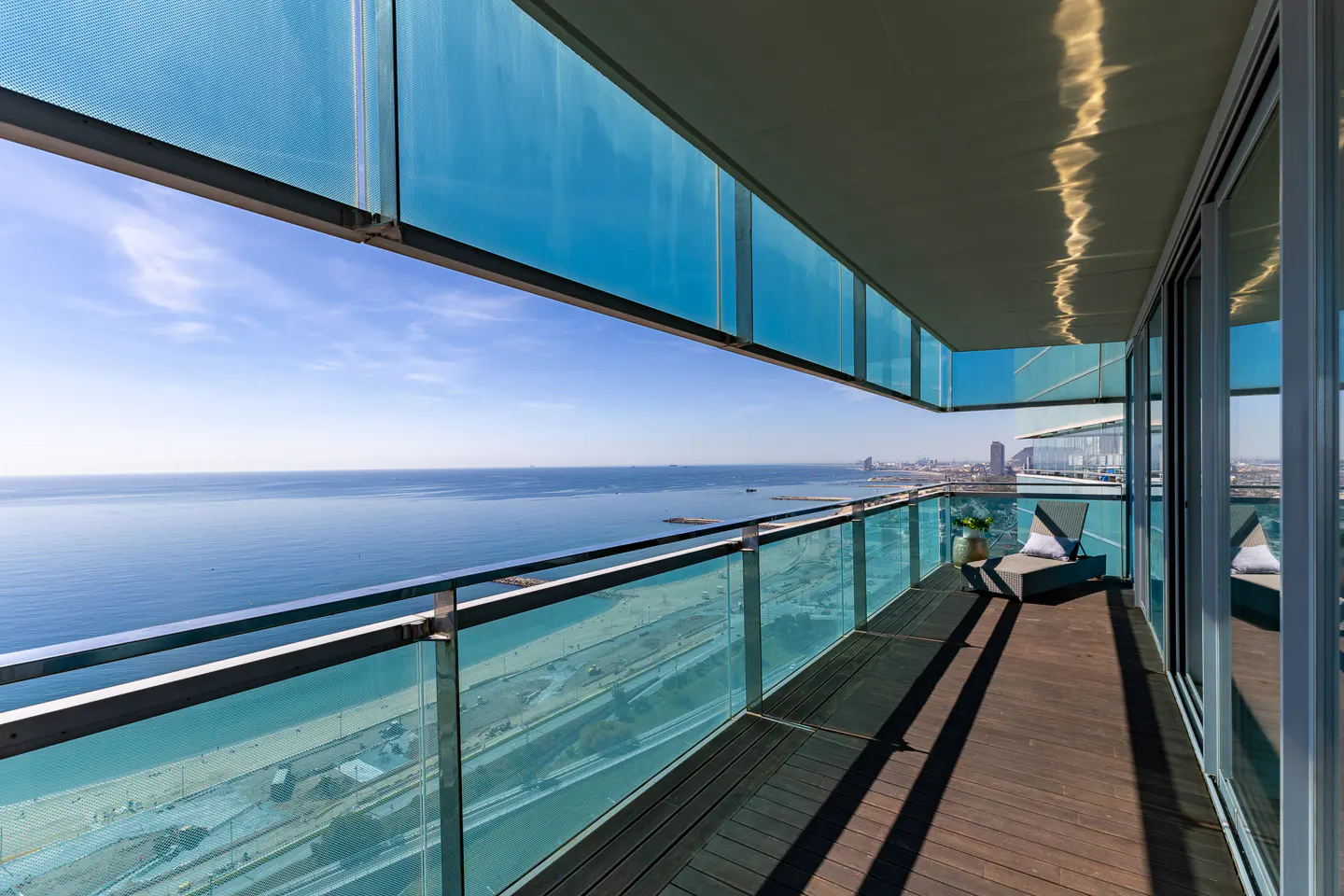 Balcony view of ocean and city. Glass railings, wood floor, and lounge chair. Blue sky and water.